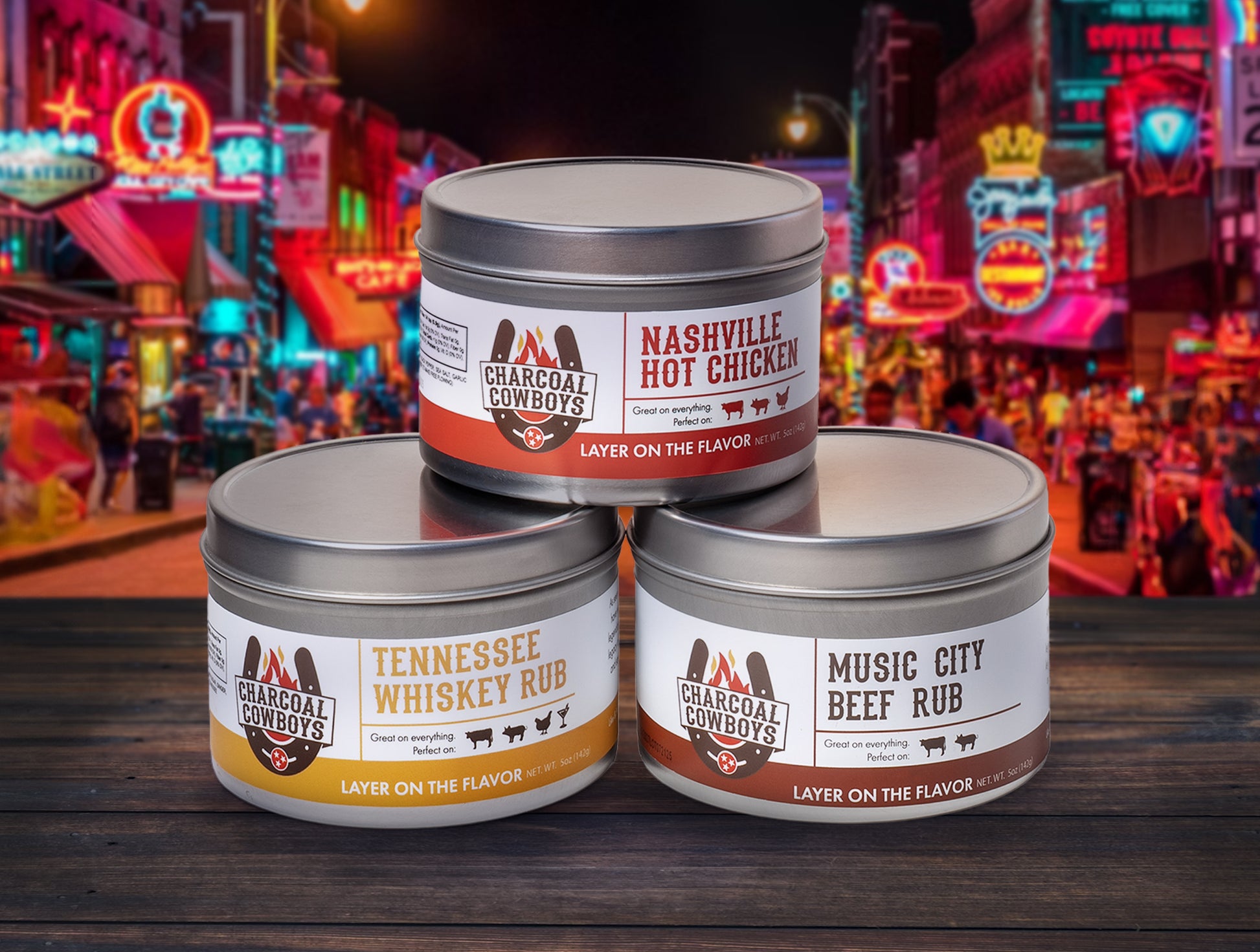 Three tins of Charcoal Cowboys seasoning with a neon-lit street scene in the background