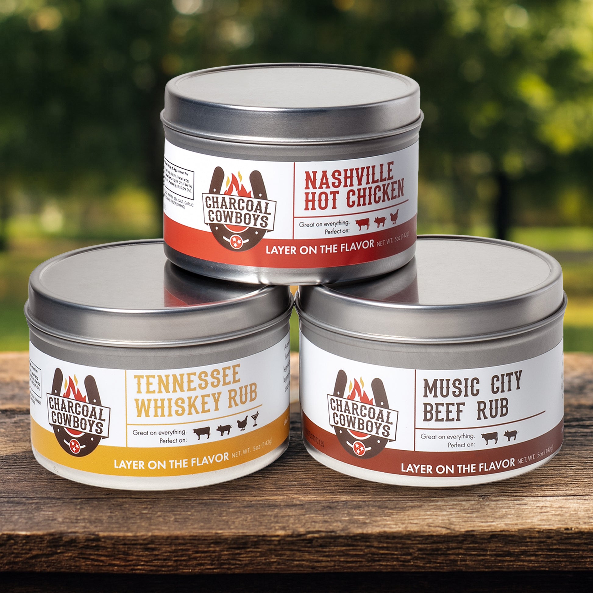 Three tins of Charcoal Cowboys seasoning on a wooden surface with a blurred green background