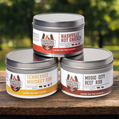 Three tins of Charcoal Cowboys seasoning on a wooden surface with a blurred green background